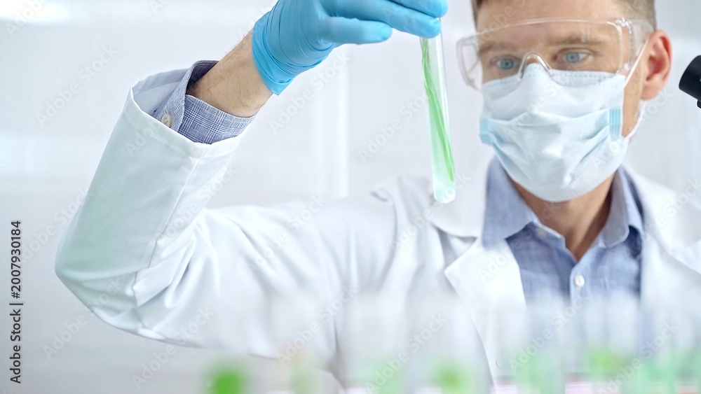 Naklejka premium Scientist with protective gloves and glasses examining lab tube with a green fluid. Microbiology and science concept