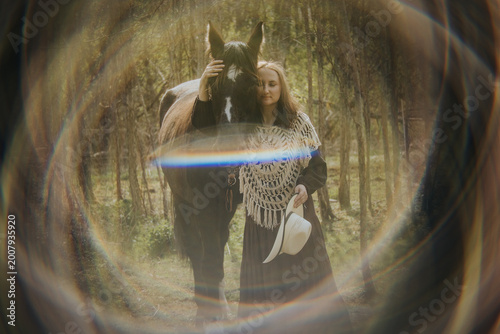 Fotografía Woman in traditional attire stands beside a black horse in a forest, wearing a d