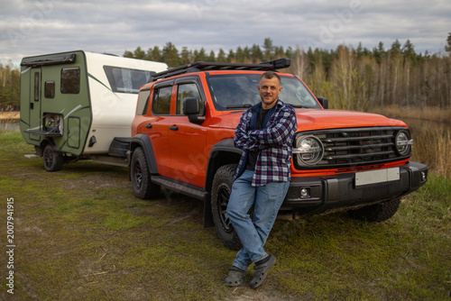 Man in a plaid shirt stands with crossed arms leaning on an orange SUV during a trip