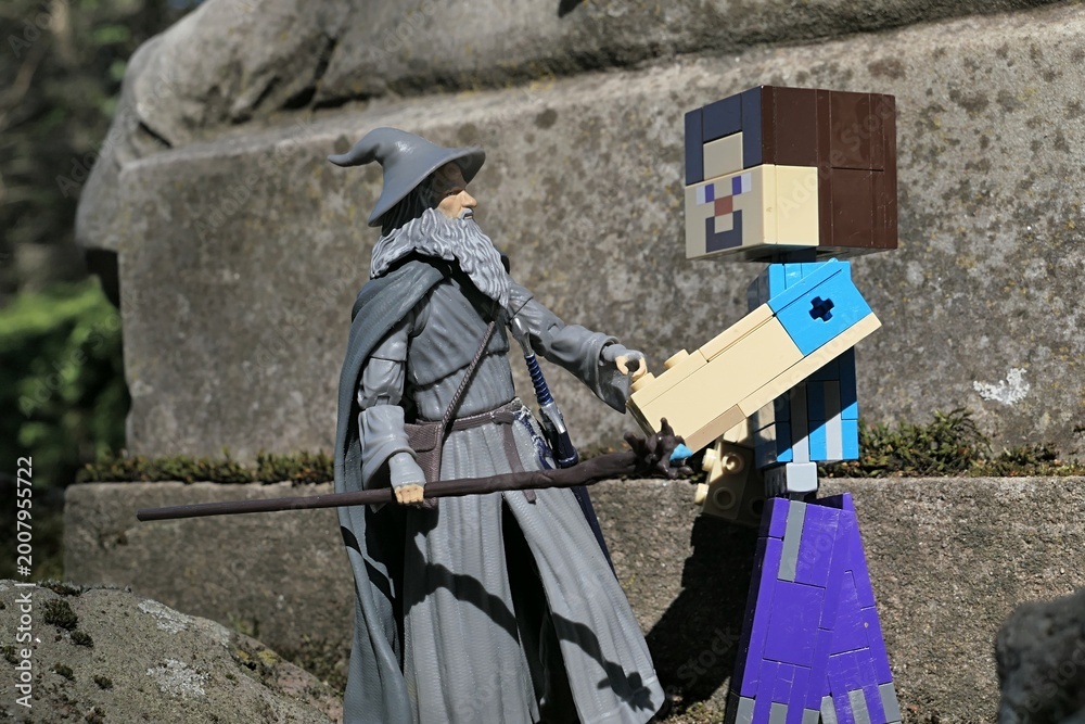 Naklejka premium Minecraft action figure of main character Steve shaking hands with figure of wizard Gandalf from Lord Of The Rings and Hobit books by J.R.R. Tolkien in front of stone pedestal, spring sunshine. 