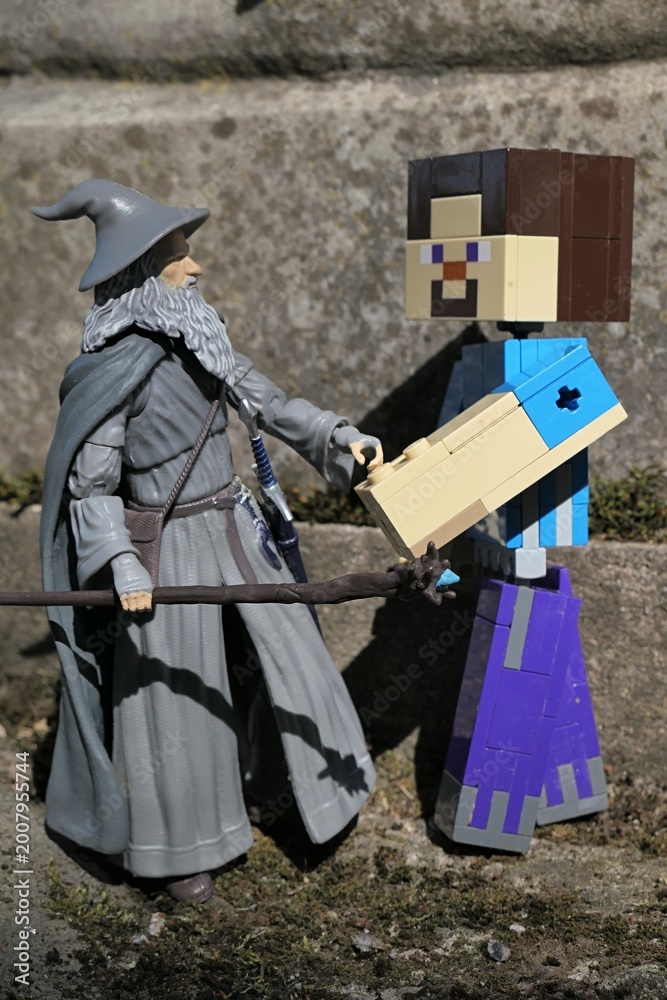 Naklejka premium Minecraft action figure of main character Steve shaking hand with action figure of wizard Gandalf from Lord Of The Rings and Hobit books by J.R.R. Tolkien in front of stone pedestal, spring sunshine. 