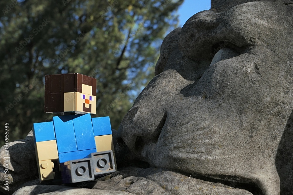 Naklejka premium Action figure of main Minecraft Character Steve looking on a face of a giant lion statue in park, spring blue skies in background. 