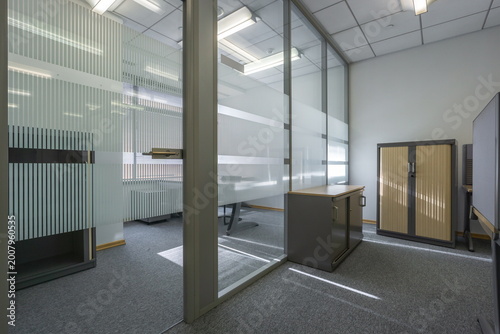 This office area features glass walls that allow natural light in, empty furniture including a desk and cabinets, creating a simple work environment in the daytime