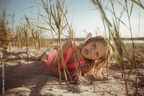 фотографія A young caucasian blonde hair girl lying on sandy beach, wearing a bright pink coral dress, joyfully shows her tongue surrounded by tall dry grass