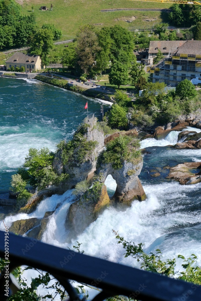 Naklejka premium Rock formation in the middle of Rhine Falls with Swiss flag, surrounded by powerful flowing water and scenic natural landscape
