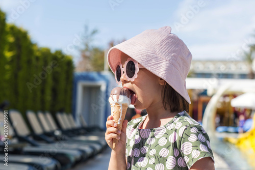 Little girl wearing pink bucket hat and stylish sunglasses eating melting ice cream cone near poolside loungers in sunny resort, summer vacation lifestyle with playful mood, childhood joy, copy space