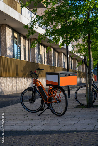 Orange Electric Delivery Bike with Cargo Box Parked on Urban Sidewalk, Netherlands. Vertical Photograph. 