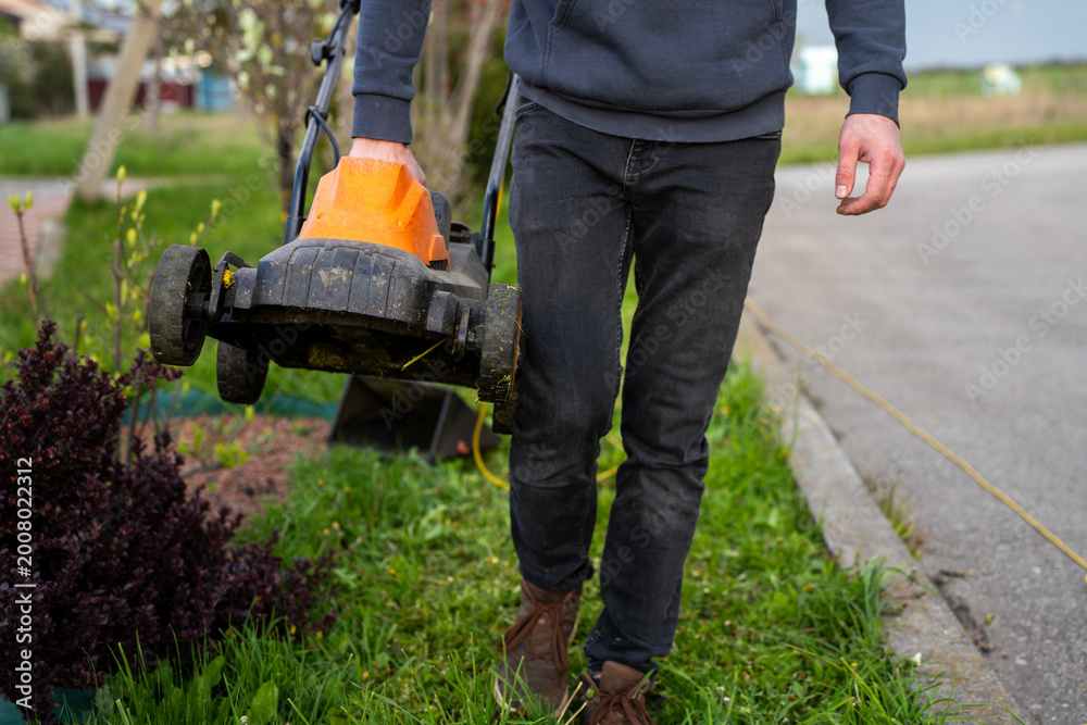 Obraz premium Person carrying electric lawn mower beside lawn and roadside, showing portable garden equipment after mowing in outdoor daylight.