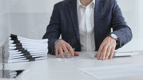 Photos Businessman sorting through stacks of documents on a white desk beside a tall pile of bound papers, symbolizing meticulous financial record keeping and bureaucracy