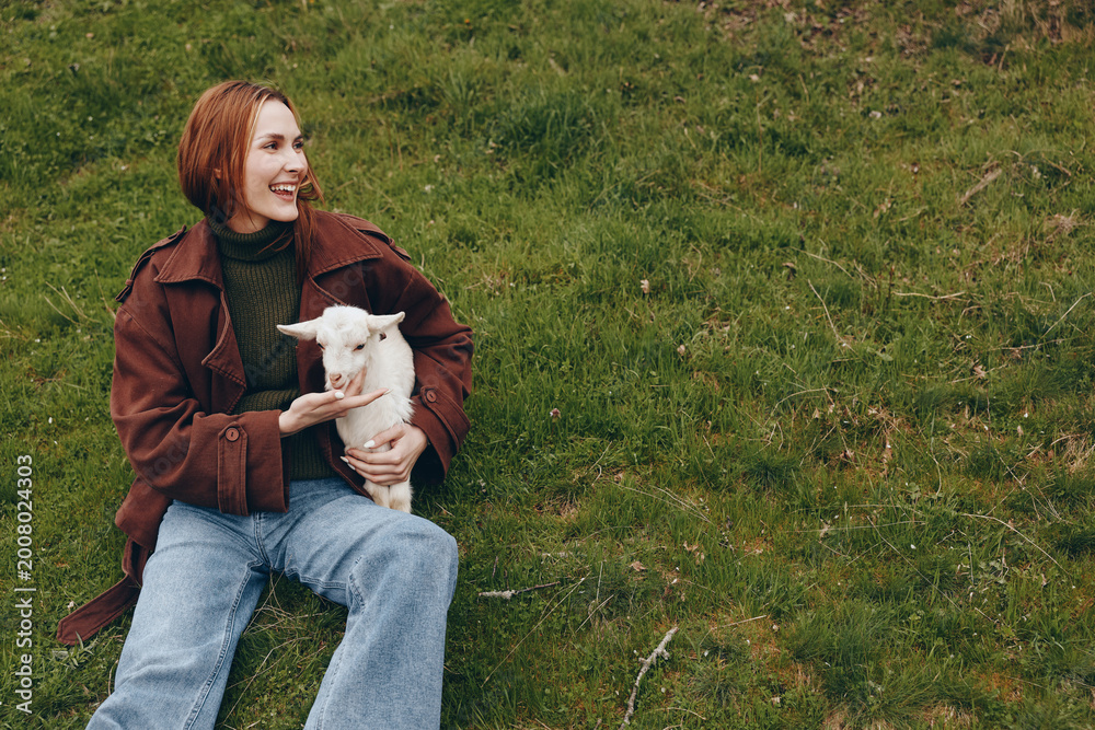 Naklejka premium Smiling woman sits on grass holding a kid goat in a park, wearing casual fashion. A warm lifestyle moment that celebrates human animal companionship outdoors, simple rural joy, and a relaxed sense of