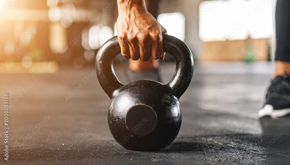 Fototapeta premium Athlete's hand gripping a black kettlebell on a gym floor, ready for strength and fitness training.