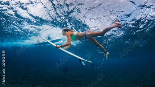 Female surfer performing duck dive underwater on surfboard beneath powerful breaking wave in tropical ocean