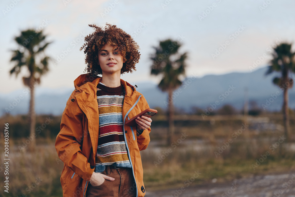 Fototapeta premium Woman lifestyle portrait outdoors, curly hair, orange jacket, striped sweater, standing on a rural road, palm trees and mountains in sunset light, relaxed travel mood