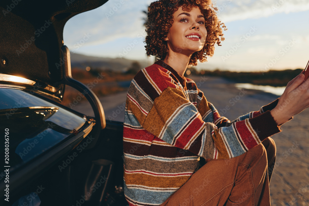 Fototapeta premium Woman lifestyle smiling at sunset while driving a car on an open road, hands on steering wheel, wearing a striped sweater, warm golden light, outdoor travel moment