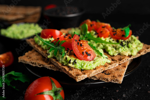 Avocado sandwich with avocado cream and rye crisp bread for snack. Fiber, fitness and diet food. Rye bread with guacamole, arugula and cherry tomatoes on dark background.