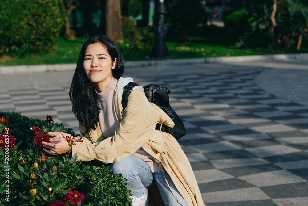 Fototapeta premium Woman kneeling beside flowers in a park, outdoor scene with backpack, casual attire, gentle smile, sunlight among greenery, calm nature moment suitable for stock photography, woman enjoying serene