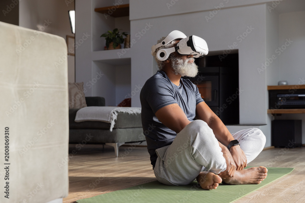 Naklejka premium Senior African American man sitting on green yoga mat in living room wearing white VR headset