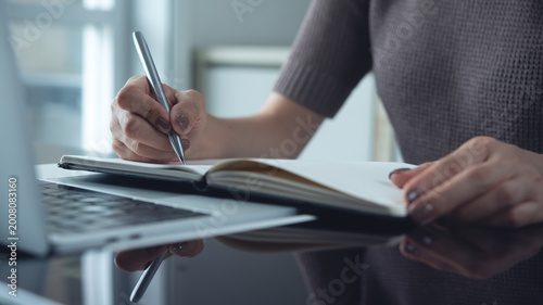 Quadro su tela Woman writing in notebook with laptop on table during working at home office, do