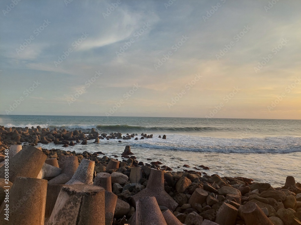 Obraz premium Golden hour seascape featuring gentle ocean waves, a rocky shoreline, and concrete breakwater structures under a dramatic cloudy sky.