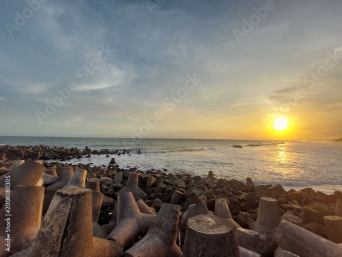Fotografie Golden hour seascape featuring gentle ocean waves, a rocky shoreline, and concrete breakwater structures under a dramatic cloudy sky