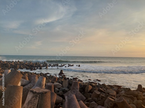 Konstfotografi Golden hour seascape featuring gentle ocean waves, a rocky shoreline, and concrete breakwater structures under a dramatic cloudy sky