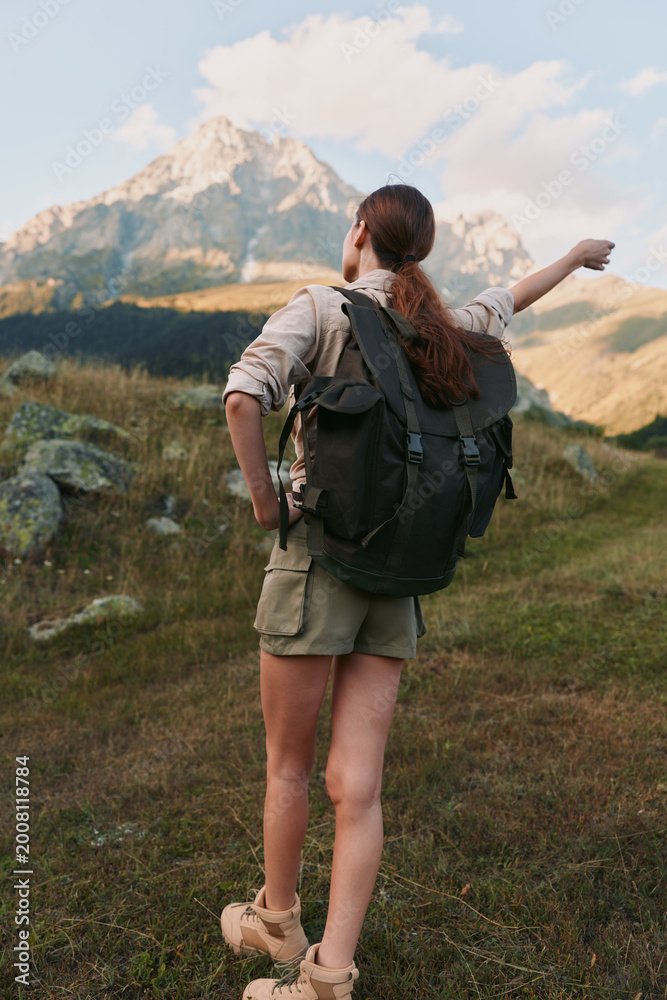 Fototapeta premium A woman hiker with a backpack stands on a grassy trail, gazing toward distant mountains. The scene conveys exploration, freedom, and the allure of outdoor adventure.