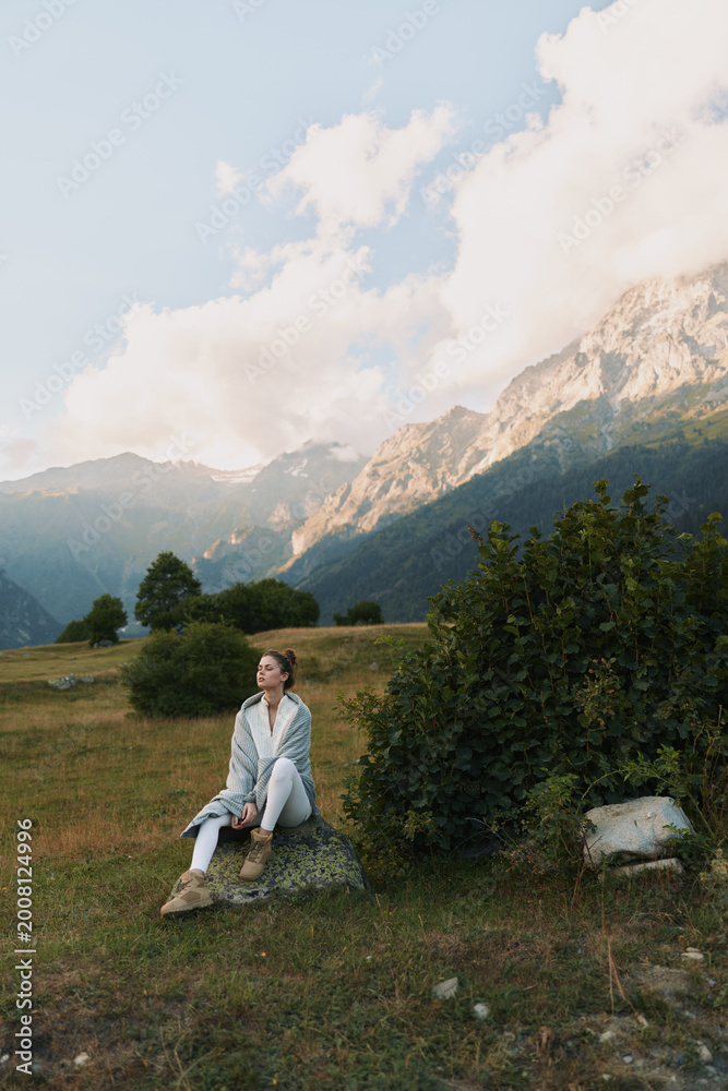 Fototapeta premium A woman sits on a rock in an open meadow with distant mountains, calm and reflective. A lifestyle portrait showing outdoor serenity and nature inspired mood.