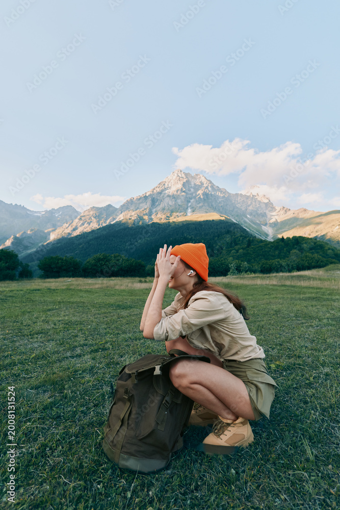 Fototapeta premium Young woman crouching in a grassy field with a backpack and orange hat, wearing casual wear. A mountain range rises in the background, conveying outdoor lifestyle and travel adventure.