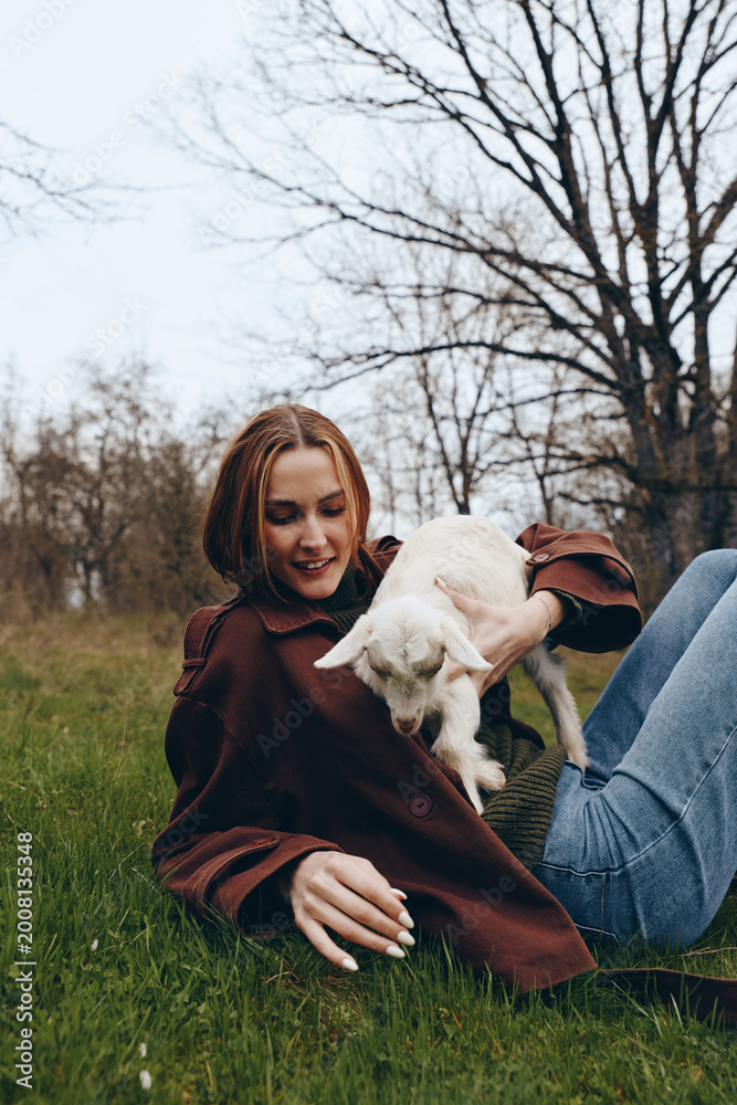 Fototapeta premium Young woman with braided hair lies on grass holding a white baby goat in her arms during a relaxed outdoor lifestyle moment in early spring.