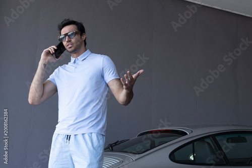 фотографія Horizontal image of a Caucasian man gesturing during a smartphone discussion near a luxury car