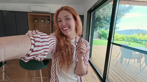 A cheerful young woman proudly displays the key to her new modern house. She beams with happiness, celebrating a significant life achievement, overlooking a beautiful garden and mountains.