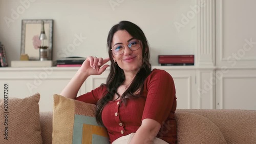 A happy young woman wearing glasses smiles and laughs while sitting comfortably on a sofa in her home, exuding confidence and a positive, relaxed vibe.