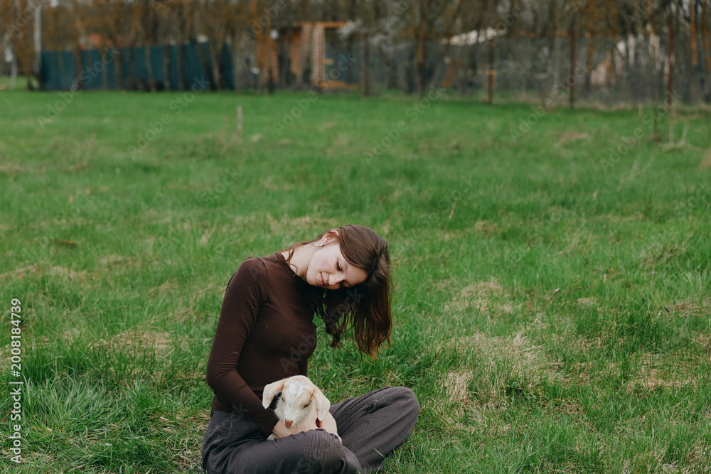 Fototapeta premium A woman in a brown sweater sits on the grass, cradling a gentle goat kid in her arms in a wide field. A calm lifestyle moment illustrating connection with animals, nature, and outdoor living.
