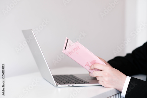 A hand holds a pink passport cover with a world map, in front of an open laptop. Clean, modern setup evoking travel planning or online booking.