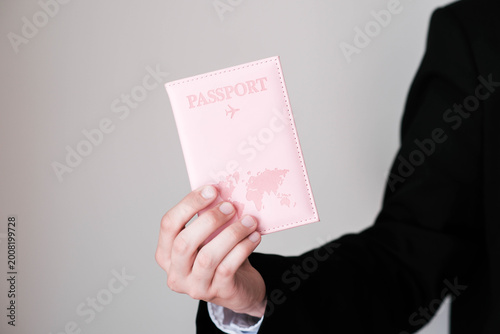 A hand in a black suit holds a stylish pink passport cover embossed text, an airplane icon, and a world map design symbolizing travel and adventure.