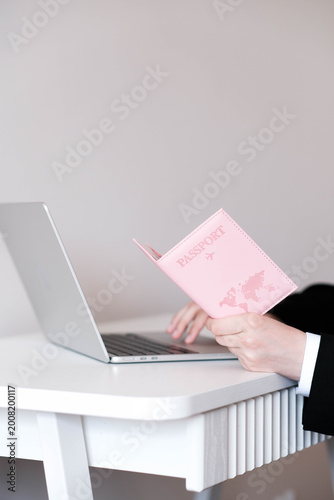 A hand holds a pink passport cover with a world map, in front of an open laptop. Clean, modern setup evoking travel planning or online booking.