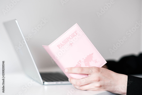 A hand holds a pink passport cover with a world map, in front of an open laptop. Clean, modern setup evoking travel planning or online booking.