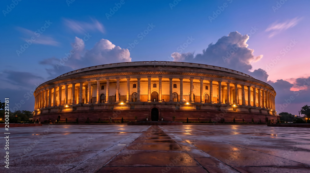 Fototapeta premium Indian Parliament Sansad Bhavan glowing with warm lights at blue hour dusk with wet plaza reflection for democracy governance and national heritage concepts