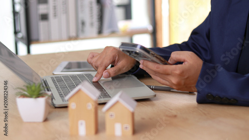 Εκτύπωση καμβά Business professional using smartphone and laptop at desk with house models symbolizing real estate investment, property management, and financial planning in a modern digital workspace environment