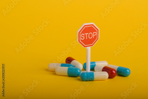 Red stop sign surrounded by colorful capsules on yellow background, representing medication restriction, drug control, healthcare regulation, or warning about pharmaceutical use.