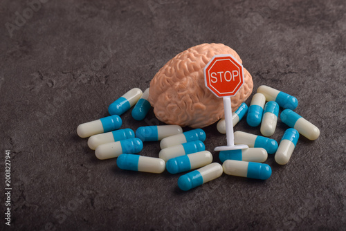 Human brain model surrounded by capsules and a stop sign on dark background, symbolizing medication control, mental health risks, drug impact on brain, or overdose warning concept.
