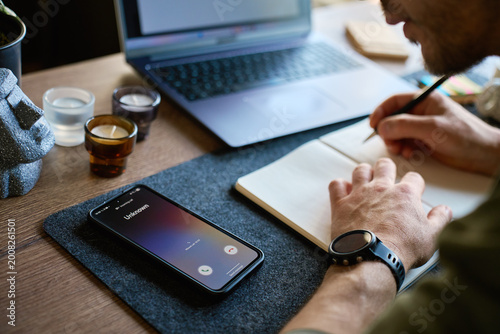 Billede på lærred Smartphone showing incoming unknown call on desk while man writes in notebook near laptop