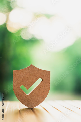 Wooden shield icon with check mark on natural bokeh background. Concept of protection, security, insurance, and guaranteed safety for business and home. High quality professional stock photo.