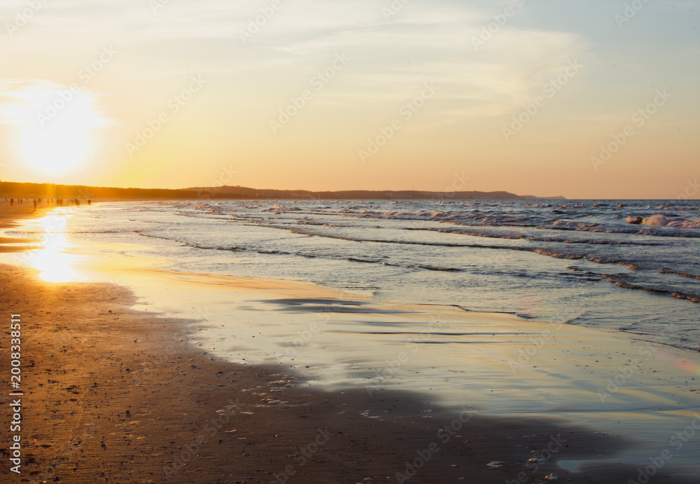 Obraz premium Gentle sea waves washing over a sandy beach during a golden sunset, with a clear sky and soft pastel colors over the Baltic Sea horizon.