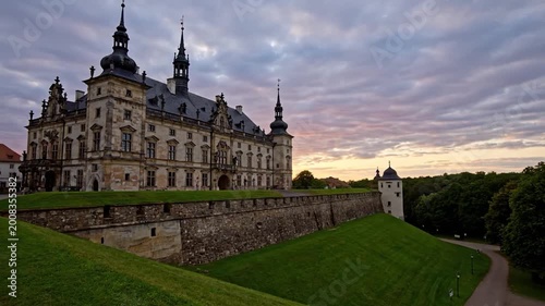 A grand, historic building with multiple towers and a long stone wall sits on a grassy hill under a colorful sunset sky