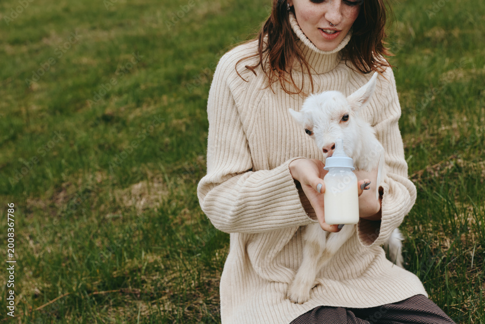 Fototapeta premium Cheerful woman outdoors, feeding a baby goat with a bottle, cozy sweater moment, gentle sunlight, peaceful field, natural colors, wholesome countryside lifestyle, calm connection