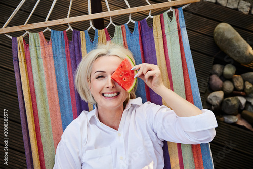 Playful mature woman holding watermelon slice over eye in hammock
