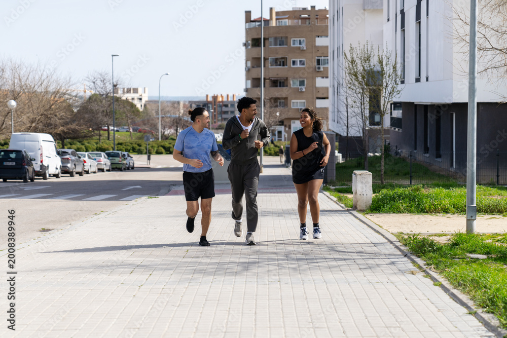Fototapeta premium Three diverse friends enjoying a group run outdoors, smiling and exercising in the city neighborhood. Fitness and healthy lifestyle