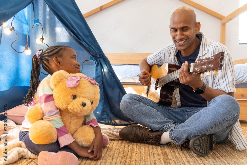 Fototapeta premium Asian father, African American child playing guitar at home holding teddy by play tent, copy space