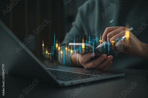 Bilde på lerret Hand using stylus on smartphone beside laptop with glowing financial chart, representing mobile trading, stock market analysis, fintech investment strategy, and digital business growth